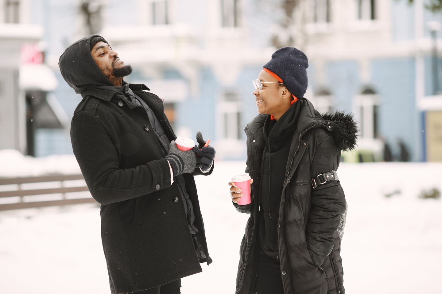 Two men in winter coats laughing and holding warm drinks on a snowy street