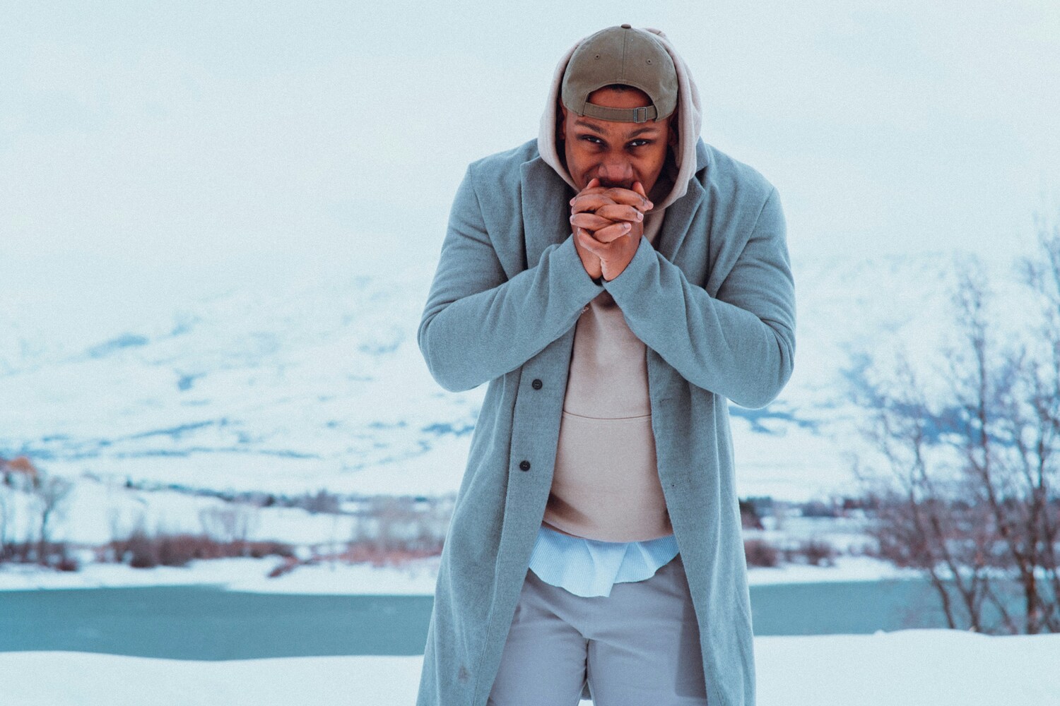 Man in a grey coat and hood warming his hands with his breath beside a snowy lake