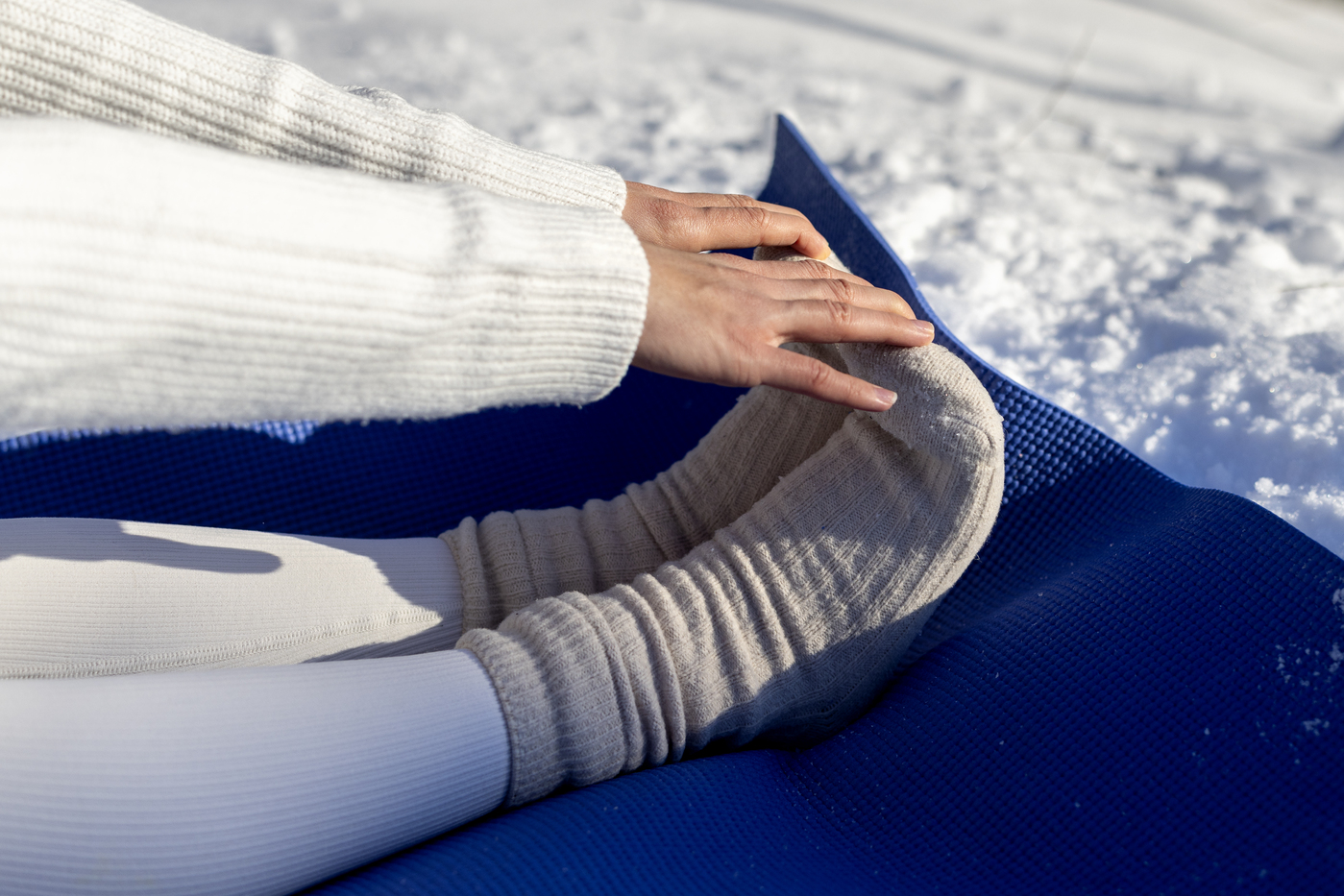 Hands in a white knitted sweater resting on knees in grey wool socks on a blue mat in the snow